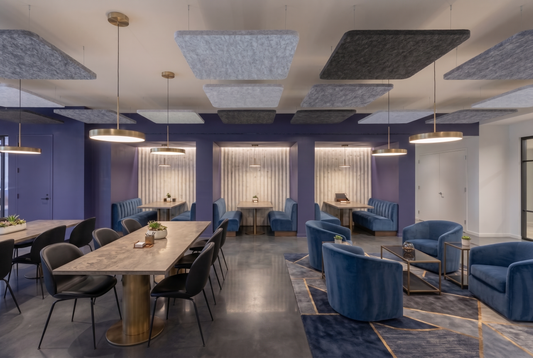Flat square PET felt acoustic rafts in grey tones hung below a standard-height ceiling in an office lounge, with a timber meeting table, blue velvet armchairs and brass pendant lights.