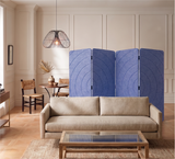 A beige fabric sofa sits in a styled living room in front of a four-panel folding screen made of the blue acoustic PET felt, with a glass-top coffee table, pendant light, and wood-paneled walls behind it.