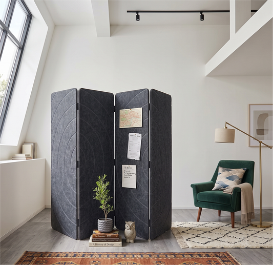A four-panel folding room divider made of dark charcoal acoustic PET felt stands in a bright, modern loft-style room. The divider is styled with a small potted plant, books, and a small owl figurine on the floor in front of it, with a green armchair, floor lamp, and patterned rug in the background.