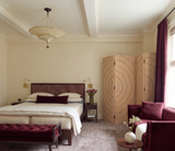 A styled bedroom interior with a bed dressed in white bedding and dark burgundy pillows, a matching upholstered bench at the foot of the bed, a burgundy armchair with a pillow, a round side table with a vase, cream walls, an acoustic PET felt folding screen, and a window with dark burgundy curtains.