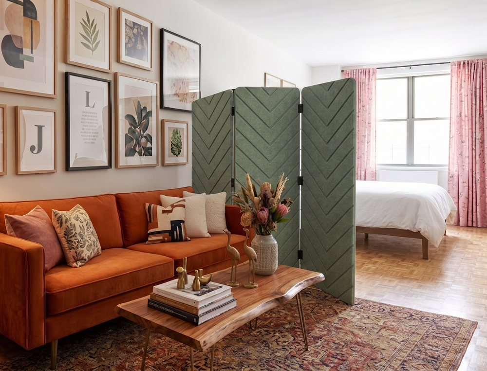 A styled interior room showing a green folding screen divider between a bright orange sofa area and a bedroom with pink curtains and a bed. The scene includes framed wall art, a wooden coffee table, books, and a rug.