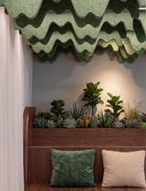A styled interior corner featuring a wooden bench with two textured throw pillows, a built-in planter with green plants, and a compound wave PET felt acoustic raft above.
