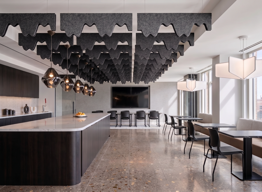 A modern open-plan office or lounge space with a long kitchen island, dining tables, black chairs, large pendant lights, and a dramatic ceiling installation of dark gray felt-like acoustic panels.