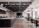 A modern open-plan office or lounge space with a long kitchen island, dining tables, black chairs, large pendant lights, and a dramatic ceiling installation of dark gray felt-like acoustic panels.