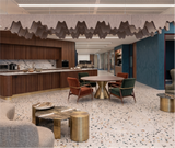 A modern interior space with a round wooden dining table, green and rust accent chairs, metallic gold side tables, and a kitchen area in the background. The room features a terrazzo-patterned floor, textured gray walls, and a distinctive wavy acoustic ceiling treatment.