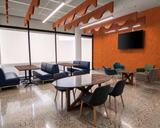 A modern office breakroom or lounge area with multiple seating options, including blue upholstered sofas, a round wooden table with dark chairs, and a bright orange hexagonal accent wall with a mounted TV.