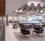 A modern interior space with a long concrete-walled corridor on the left and a lounge/cafeteria area on the right, featuring wavy ceiling panels, tables, chairs, and a textured rug.