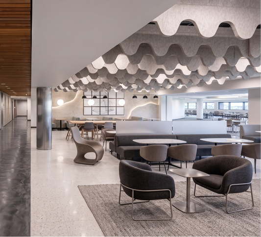 A modern interior space with a long concrete-walled corridor on the left and a lounge/cafeteria area on the right, featuring wavy ceiling panels, tables, chairs, and a textured rug.