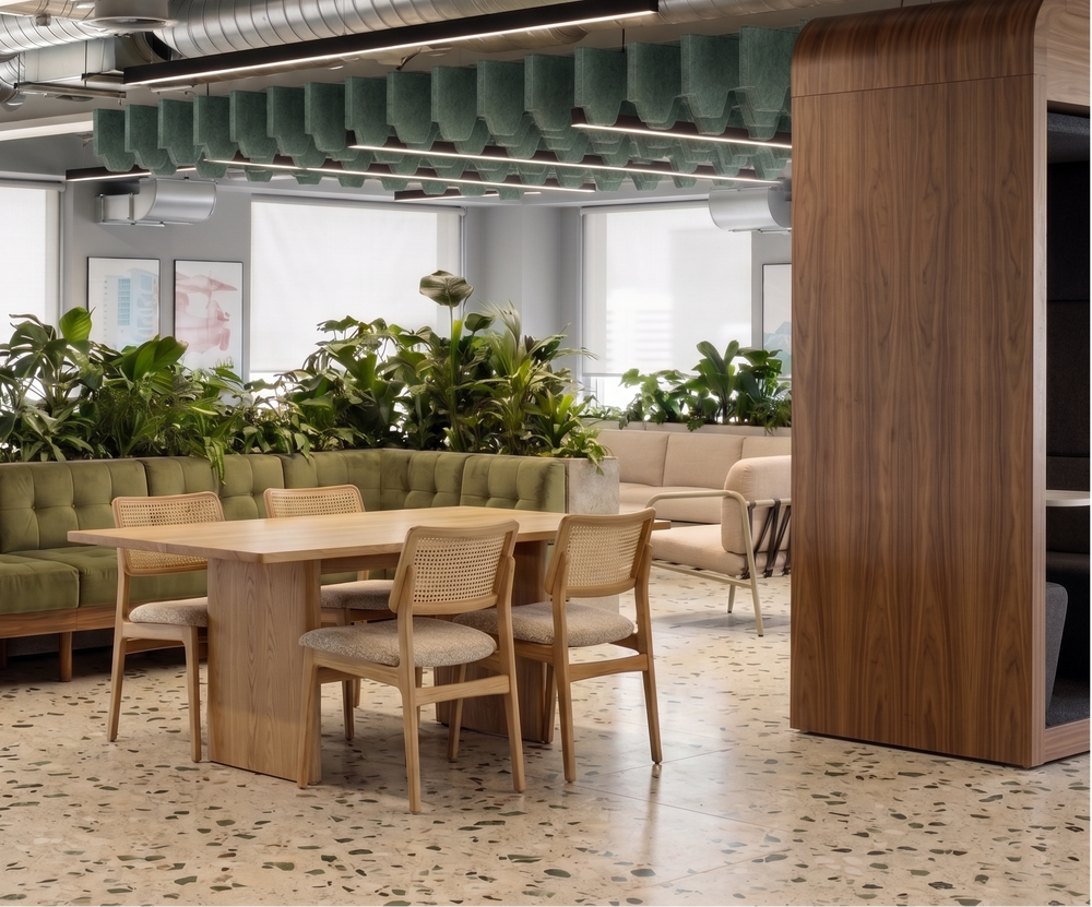 A modern office interior featuring a wooden dining table and four upholstered chairs in the foreground, with a long green tufted bench and multiple green plants behind it. The space has exposed ceiling ducts, bright windows, and a wood-paneled column on the right.