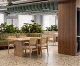 A modern office interior featuring a wooden dining table and four upholstered chairs in the foreground, with a long green tufted bench and multiple green plants behind it. The space has exposed ceiling ducts, bright windows, and a wood-paneled column on the right.