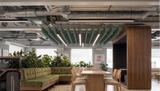 An interior office or lounge space with exposed industrial ceiling ducts, a green tufted sectional sofa, a wooden dining table and chairs, and a large wood partition wall. The scene is well lit with natural light from windows and features a modern, open layout.