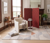 A styled living room scene featuring a prominent red folding screen with concentric circular patterns, set next to a cream armchair and a geometric patterned rug on a light wood floor.