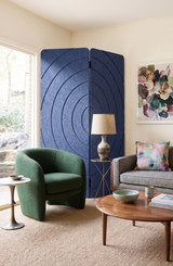 A styled living room featuring a prominent dark blue circular-patterned folding screen behind a green armchair and a gray sofa with a floral print pillow.