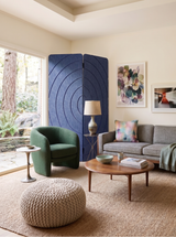 A styled living room interior featuring a dark green armchair, a round knitted pouf, a gray sofa, a wooden coffee table, and a tall blue folding screen against a cream wall.