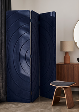 A three-panel folding screen with a deep navy blue circular concentric pattern, styled in a modern room next to a wooden cabinet, lamp, and stool.