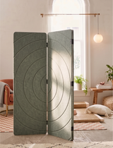 A three-panel folding room divider with a textured, circular concentric pattern in a muted sage green color, styled in a bright, modern room with a window, pendant light, and cozy decor.