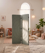 A three-panel folding room divider with a textured circular concentric pattern in sage green, styled in a bright, modern living room.