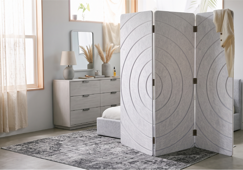 A three-panel folding room divider with a textured light gray fabric and concentric circle pattern, styled in a bright, modern bedroom next to a bed and dresser.