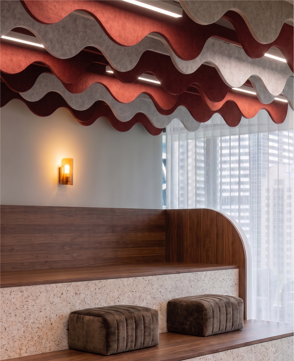 Modern indoor seating area with tiered wooden benches, textured terrazzo-look side panels, two dark brown velvet ottomans, a wall sconce, and wavy red and gray acoustic ceiling panels overhead.