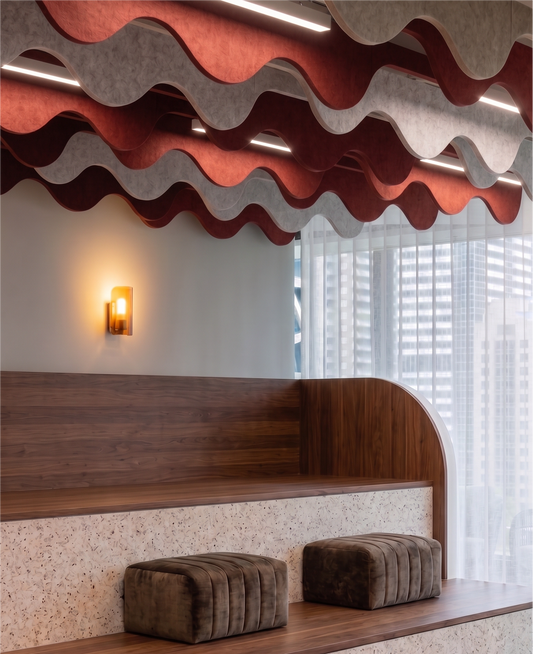 Modern indoor seating area with tiered wooden benches, textured terrazzo-look side panels, two dark brown velvet ottomans, a wall sconce, and wavy red and gray acoustic ceiling panels overhead.
