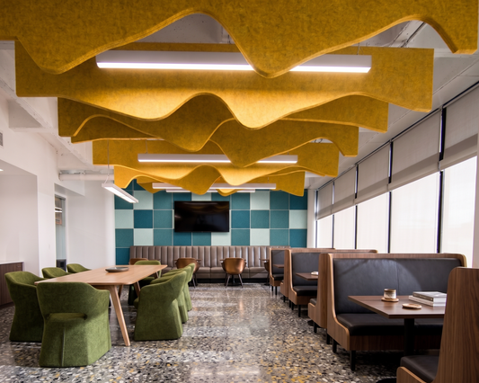 A modern office lounge or breakroom interior with wavy yellow acoustic ceiling panels, green upholstered chairs, wooden tables, booth seating, and a blue-green checkered accent wall in the background.