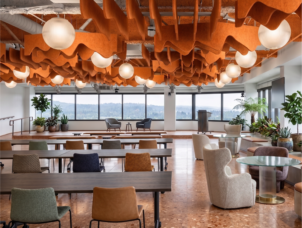 A modern, spacious office or lounge interior with rows of wooden desks and chairs, round pendant lights, and a prominent orange wavy ceiling installation. Large windows look out onto a distant landscape, and several potted plants are placed throughout the space.