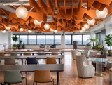 A modern, spacious office or lounge interior with rows of wooden desks and chairs, round pendant lights, and a prominent orange wavy ceiling installation. Large windows look out onto a distant landscape, and several potted plants are placed throughout the space.