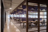 A modern open-plan office interior with rows of desks, office chairs, purple acoustic wall panels, and large wood-framed glass dividers. The space has a polished concrete floor, a patterned gray rug, and large windows in the background.